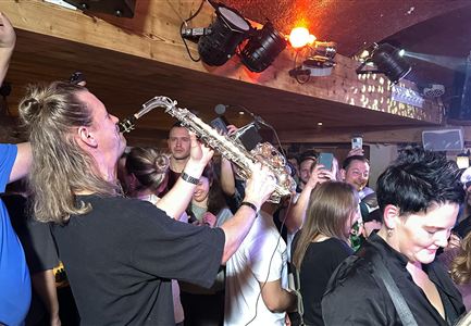 Un vivace ambiente di festa con un sassofonista che suona per la folla.  
Le persone ballano e godono della musica in un'atmosfera festosa.