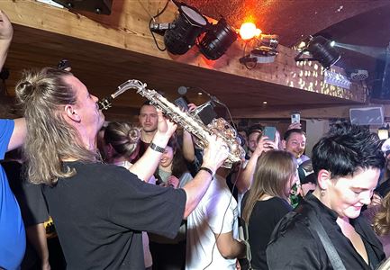 Ein lebhaftes Partyambiente mit einem Saxophonisten, der für die Menge spielt. Menschen tanzen und genießen die Musik in einer festlichen Atmosphäre.