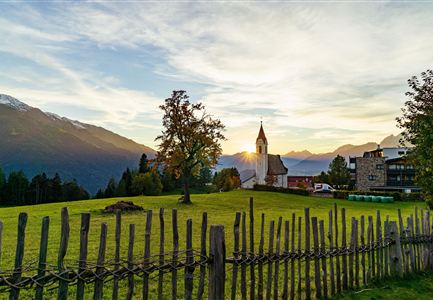 Een pittoresk landschap met een kerk en een hek van houten planken. Op de achtergrond zijn bergen en een zonsondergang te zien.