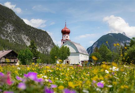 Eine malerische Kirche mit einer roten Kuppel steht inmitten eines bunten Blumenfeldes. Im Hintergrund sind majestätische Berge und ein blauer Himmel sichtbar.