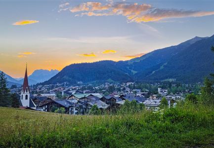 Eine malerische Aussicht auf ein Dorf in den Bergen, umgeben von grünen Wiesen und hohen Bergen. Der Himmel ist in sanften Farben erleuchtet, während die Kirchturmspitze ins Bild ragt.