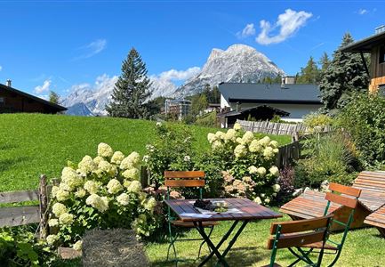 Ein schöner Garten mit blühenden Pflanzen und gemütlichen Sitzgelegenheiten. Im Hintergrund sieht man beeindruckende Berge und einen blauen Himmel.