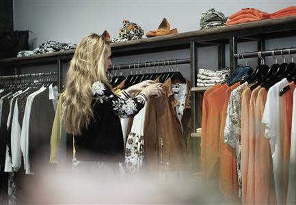 A woman is standing in a clothing store and is looking at clothes. Next to her, various tops and blouses in warm colors are displayed.