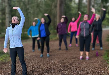 A group of people is exercising outdoors on a forest path. The participants are raising their arms and wearing colorful sportswear.