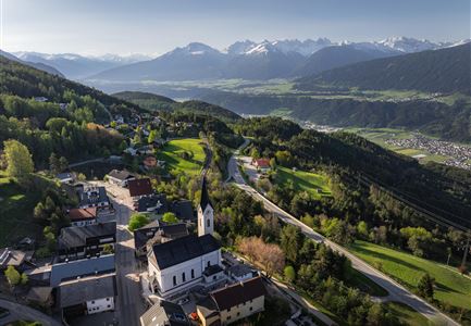 Eine malerische Landschaft mit einem kleinen Dorf und einer Kirche. Im Hintergrund sind die Alpen und grünes, sanft gewelltes Land zu sehen.
