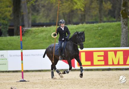 Eine Reiterin in schwarzer Kleidung reitet auf einem schwarzen Pferd in einer Reithalle. Sie führt eine Übung mit einem langen Stock und einer Hütchenmarkierung durch.