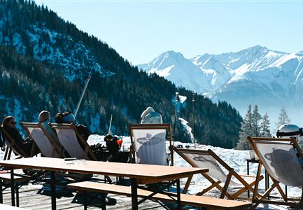 Eine gemütliche Sitzgruppe auf einer Terrasse mit Blick auf schneebedeckte Berge. Die Sonne scheint und es ist eine klare Winterlandschaft zu sehen.