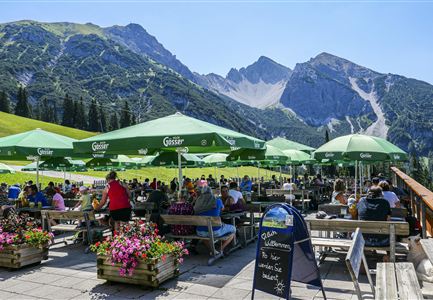 Eine schöne Terrasse mit vielen Leuten und grünen Sonnenschirmen. Im Hintergrund sind beeindruckende Berge zu sehen.