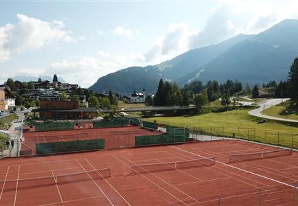 Ein Tennisplatz mit roter Erde, umgeben von Bergen und Bäumen. Im Hintergrund sind einige Gebäude und eine Straße sichtbar.