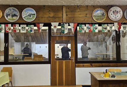 Ein gemütlicher Raum mit Holzmöbeln und dekorativen Wandtellern. Im Hintergrund sind zwei Personen sichtbar, die an einem Tisch stehen.