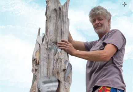 Ein Künstler arbeitet an einer Holzskulptur mit Herzmotiven. Der Himmel ist blau mit einigen Wolken.