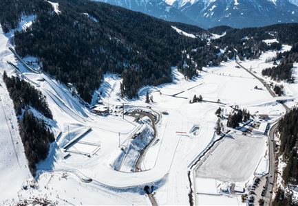 Eine malerische Schneelandschaft mit bewaldeten Hügeln und einer Skipiste. Im Hintergrund sind schneebedeckte Berge und ein klarer Himmel zu sehen.