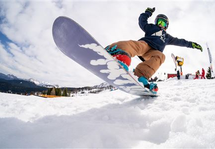Ein Snowboardfahrer springt in den Schnee. Im Hintergrund sind Berge und andere Menschen zu sehen.