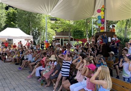 Een grote menigte zit onder een zonnescherm op een festivalterrein. Veel kinderen en volwassenen kijken nieuwsgierig naar een voorstelling.