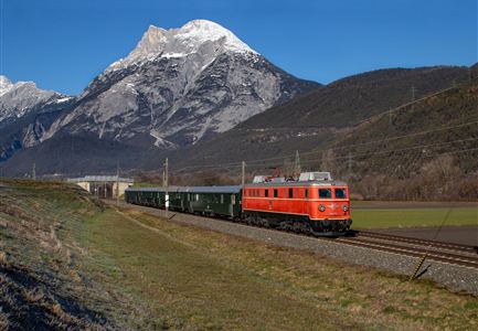 Ein roter Zug fährt entlang einer malerischen Landschaft mit beeindruckenden Bergen im Hintergrund. Der Himmel ist klar und strahlend blau.