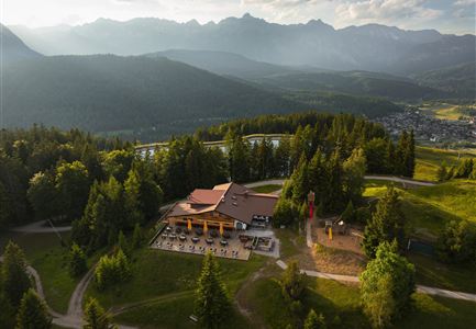 A mountain landscape with a modern building in the center, surrounded by trees. In the background, majestic mountains and an expansive sky can be seen.