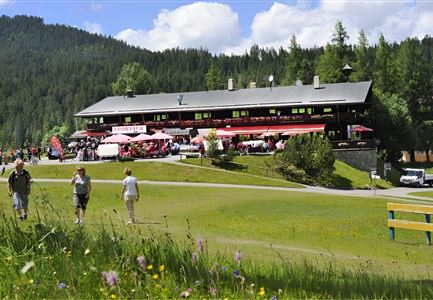 Een groot restaurant in een groene omgeving met tafels buiten. Mensen wandelen voorbij en genieten van het mooie weer.