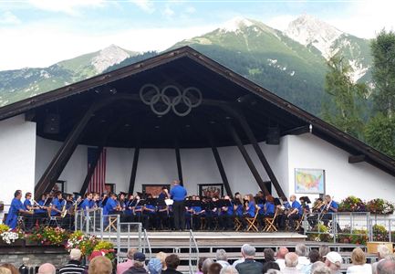 Eine Musikgruppe tritt auf einer Bühne auf, umgeben von Bergen. Das Publikum sitzt vor der Bühne und genießt die Aufführung.