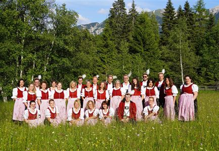 Eine Gruppe von Menschen in traditionellen Trachten steht auf einer Wiese umgeben von Bäumen. Im Hintergrund sind Berge und ein blauer Himmel sichtbar.