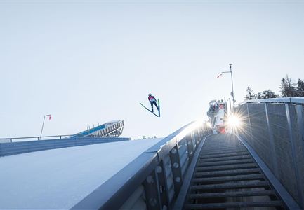 Ein Skispringer in der Luft über einer Sprungschanze. Die Szene wird von hellem Sonnenlicht erhellt.