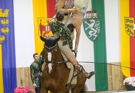 Eine elegante Pferdetricks, bei dem eine Darstellerin auf dem Pferd steht und eine akrobatische Pose einnimmt. Im Hintergrund sind Fahnen und Blumenarrangements zu sehen.
