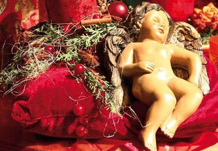 A festive decoration featuring a sleeping angel figure on a red cushion. Surrounded by Christmas accessories and pine branches.