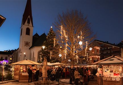 Ein festlich geschmückter Weihnachtsmarkt mit vielen Lichtern und Ständen. Im Hintergrund ist eine Kirche und ein großer, beleuchteter Baum zu sehen.
