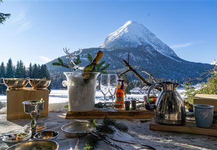 Ein schön gedeckter Tisch im Schnee mit Blick auf einen Berg. Gläser und Getränke sind bereit, um die winterliche Landschaft zu genießen.