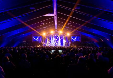 A large audience at a concert in a hall. Brightly lit stage with colorful lights and a large spectator area.