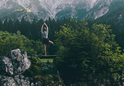 Eine Person macht Yoga auf einem Steinfeld in der Natur. Im Hintergrund sind majestätische Berge und grüne Wälder zu sehen.