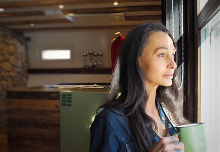 Eine Frau steht am Fenster und blickt nachdenklich nach draußen. Sie hält eine Tasse in der Hand und scheint einen Moment der Ruhe zu genießen.