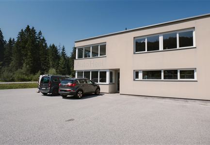 Ein modernes Gebäude mit großen Fenstern und einem Parkplatz. Zwei Autos stehen vor dem Gebäude, umgeben von Bäumen.