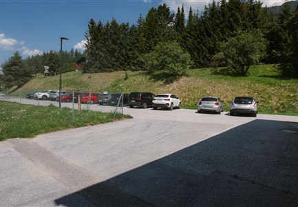 A parking lot with several cars, surrounded by trees and a green meadow. In the background, the sky is partly cloudy.