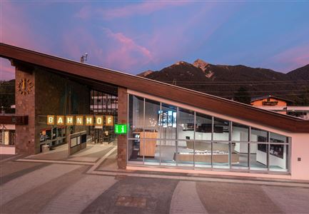 A modern train station with a slanted roof structure and large windows. In the background, mountains and a colorful evening sky are visible.
