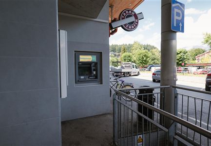 An ATM is located in a modern environment. In the background, cars and trees can be seen.