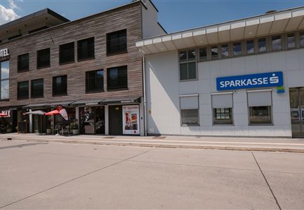 A modern building with wooden cladding and a bright bank counter of the savings bank can be seen. The street is quiet and provides ample space for pedestrians.