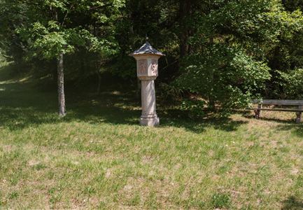 Ein schöner, grüner Park mit einem Vogelhaus auf einem Säulenstand. Im Hintergrund sind Bäume und eine Holzbank zu sehen.