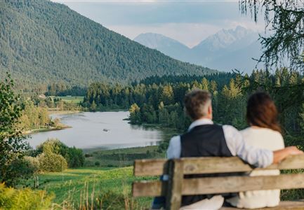 Ein Paar sitzt auf einer Bank und blickt auf einen malerischen See, umgeben von Bergen und Bäumen. Die Landschaft wirkt ruhig und idyllisch.