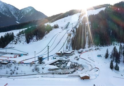 Un paesaggio montano invernale con pendii coperti di neve e impianti di risalita. In primo piano si vede una piccola stazione sciistica con parcheggi e alberi circostanti.