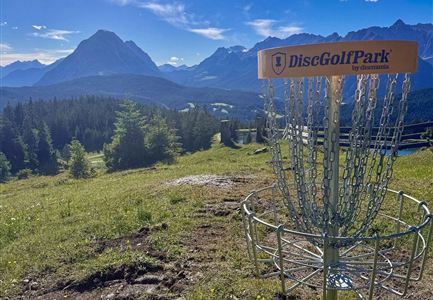 Ein Discgolf-Korb steht auf einer Wiese mit einer atemberaubenden Berglandschaft im Hintergrund. Der Himmel ist blau mit einigen Wolken.