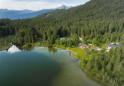 Ein malerischer See umgeben von dichtem Wald und sanften Hügeln. Im Hintergrund sind Berge und eine ruhige, klare Wasseroberfläche zu sehen.