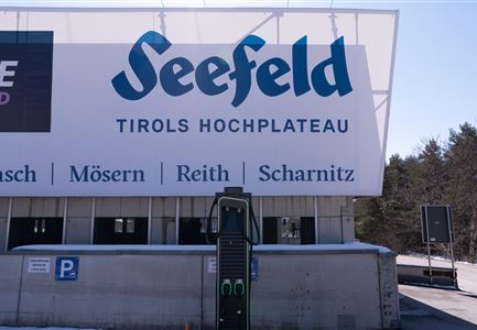 Ein großes Schild für Seefeld, Tirols Hochplateau, steht im Vordergrund. Im Vordergrund befindet sich eine Ladestation für Elektrofahrzeuge.