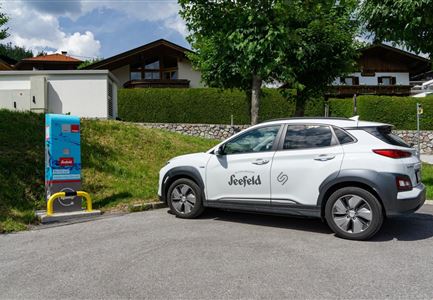 An electric car is parked at a charging station in a quiet environment. In the background, green trees and buildings can be seen.