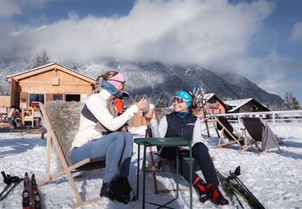 Zwei Frauen sitzen entspannt auf einer Terrasse im Schnee. Im Hintergrund sind Hütten und verschneite Berge zu sehen.