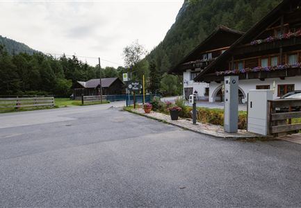 A quiet rural street with a traditional building and green spaces. In the foreground, some flower pots and a charging station for electric cars can be seen.