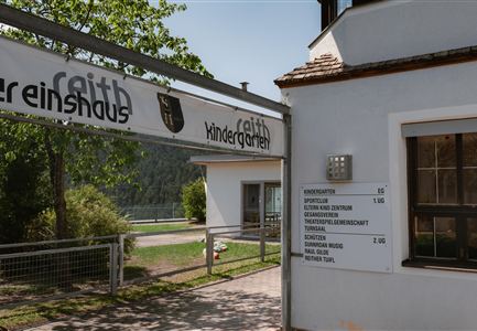 An entrance to a kindergarten with a sign and an information area. In the background, trees and a green environment are visible.