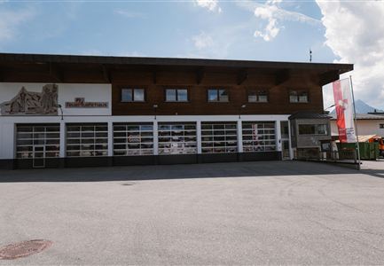 Ein modernes Gebäude mit Holzverkleidung und großen Fenstern. Der Bereich vor dem Gebäude ist asphaltiert und es gibt eine Fahne an der Seite.
