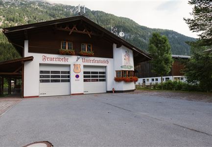 Ein traditionelles Feuerwehrhaus mit Holzfassade und zwei großen Garagentoren. Im Hintergrund sind Berge und Bäume zu sehen.