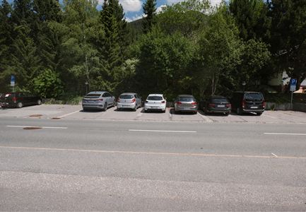 A parking lot with several cars, surrounded by trees. The road is visible directly next to the parking lot.