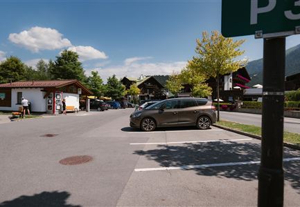 A parking lot with a gray car and surrounding buildings in alpine style. In the background, trees and a blue sky are visible.